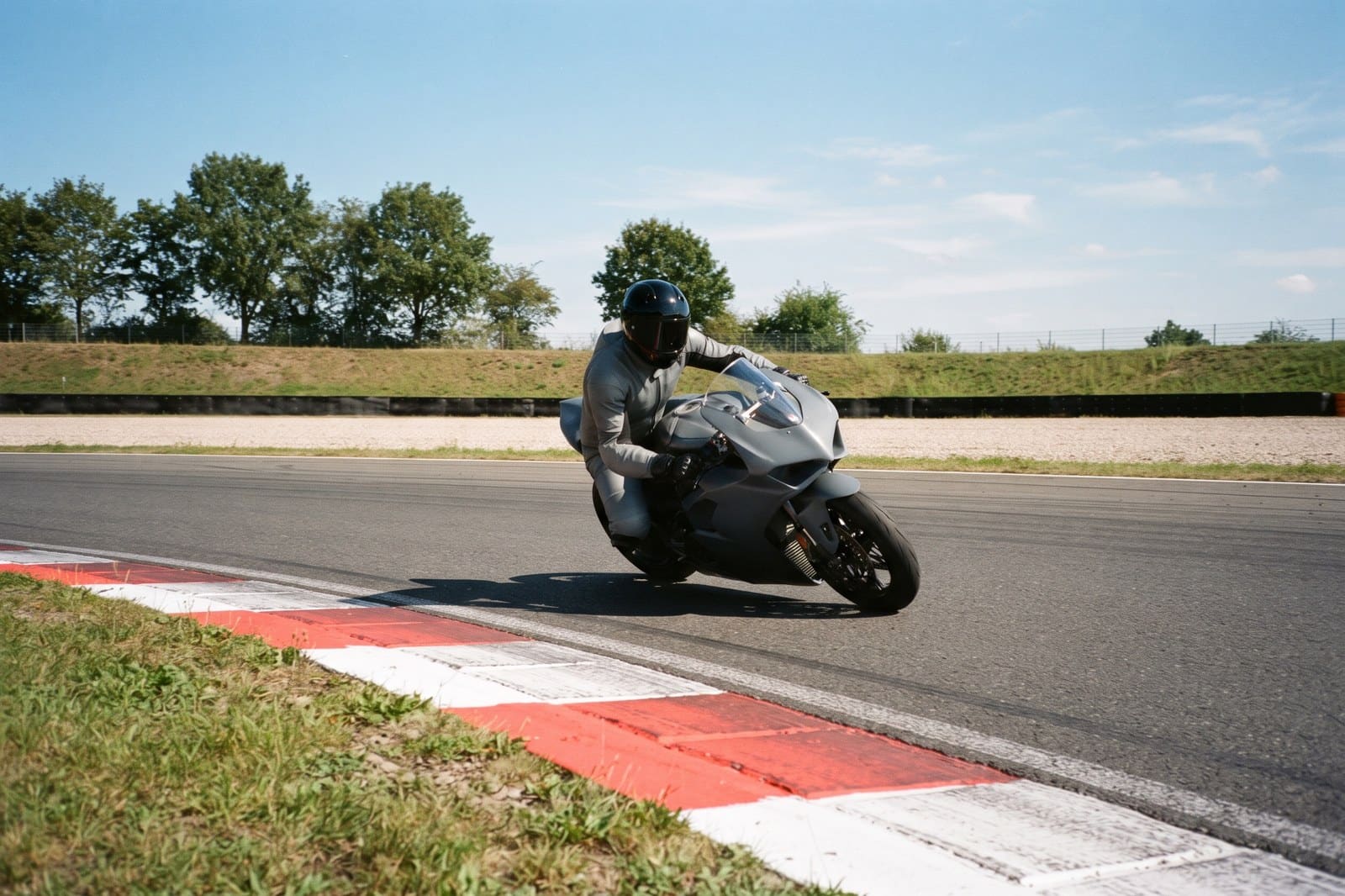 Motorradfahrer in grauer Lederkombi lehnt ein Sportmotorrad in eine Kurve auf der Rennstrecke, rot-weiße Curbs und Sonnenschein, Unfallversicherung Trackday